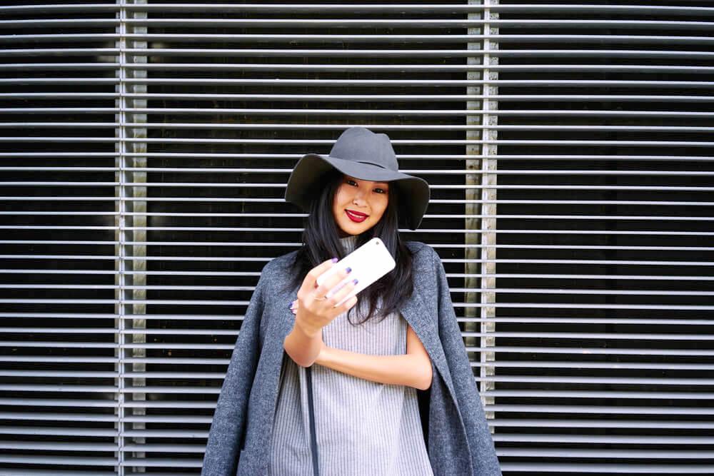 Fashionable young woman in hat and grey coat taking a selfie